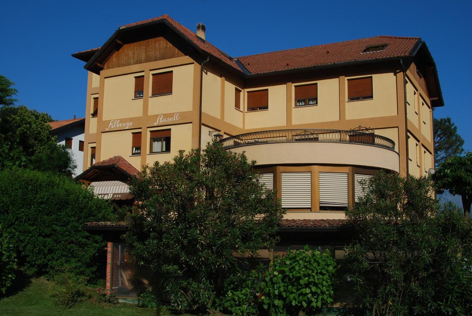 Albergo Rusall – hotel galben cu balcon curbat si veranda, inconjurat de vegetatie mediteraneana, Tremezzo Albergo Rusall – hotel galben cu balcon curbat si veranda, inconjurat de vegetatie mediteraneana, Tremezzo