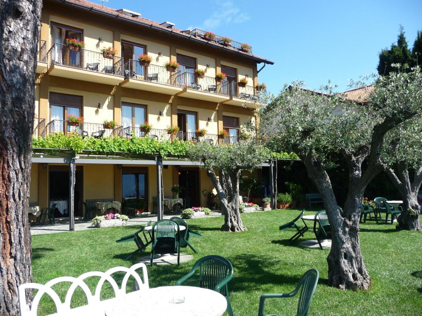 Albergo Rusall – fatada hotelului cu balcoane ornate cu muscate rosii si gradina cu maslin si scaune verzi, Tremezzo Albergo Rusall – fatada hotelului cu balcoane ornate cu muscate rosii si gradina cu maslin si scaune verzi, Tremezzo