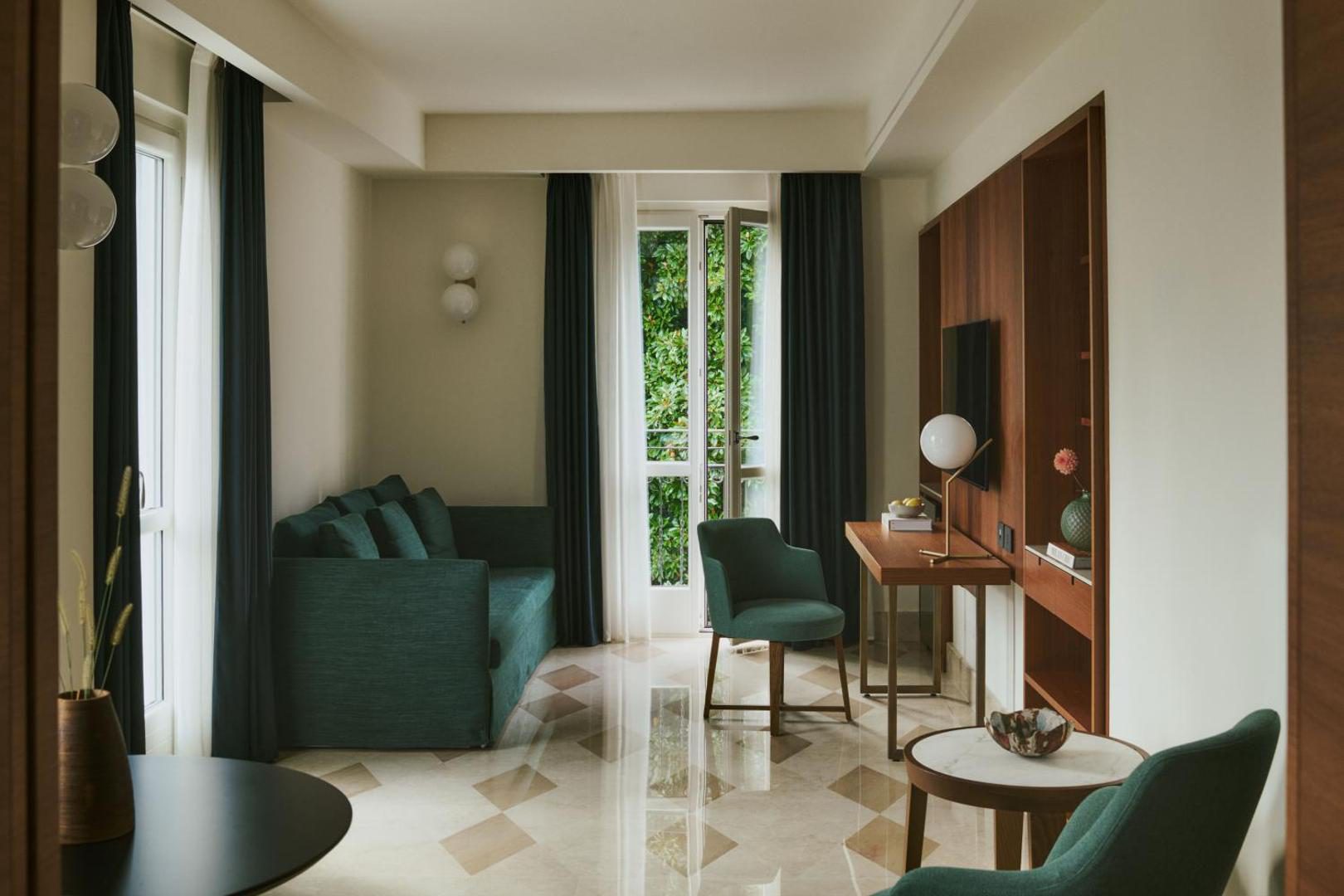 Hotel Belvedere – camera renovata cu canapea verde, birou si usi spre terasa cu vegetatie Hotel Belvedere – camera renovata cu canapea verde, birou si usi spre terasa cu vegetatie