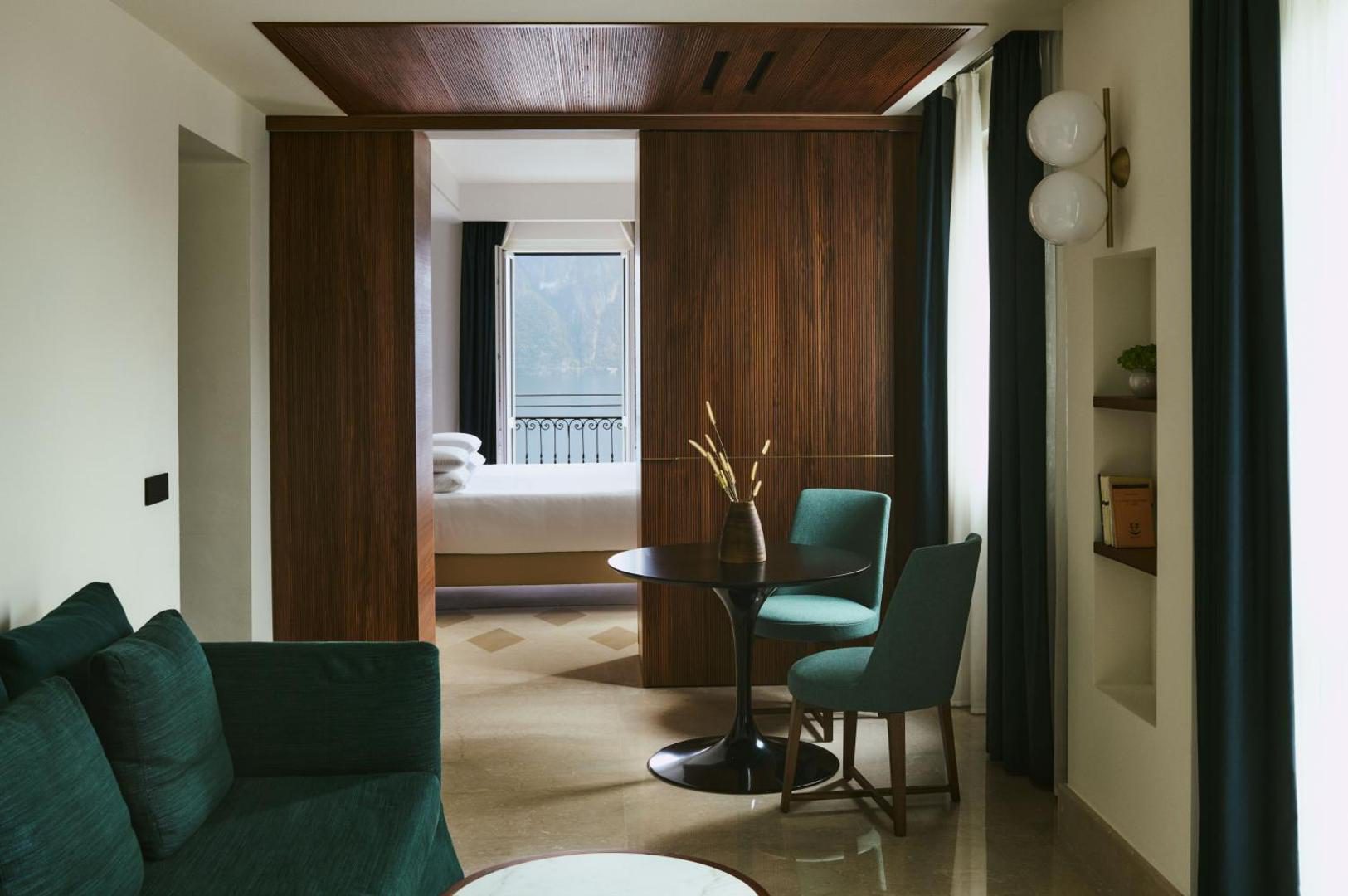 Hotel Belvedere – zona de living a suitei cu canapea verde, masa rotunda si vedere spre dormitor cu lac Hotel Belvedere – zona de living a suitei cu canapea verde, masa rotunda si vedere spre dormitor cu lac