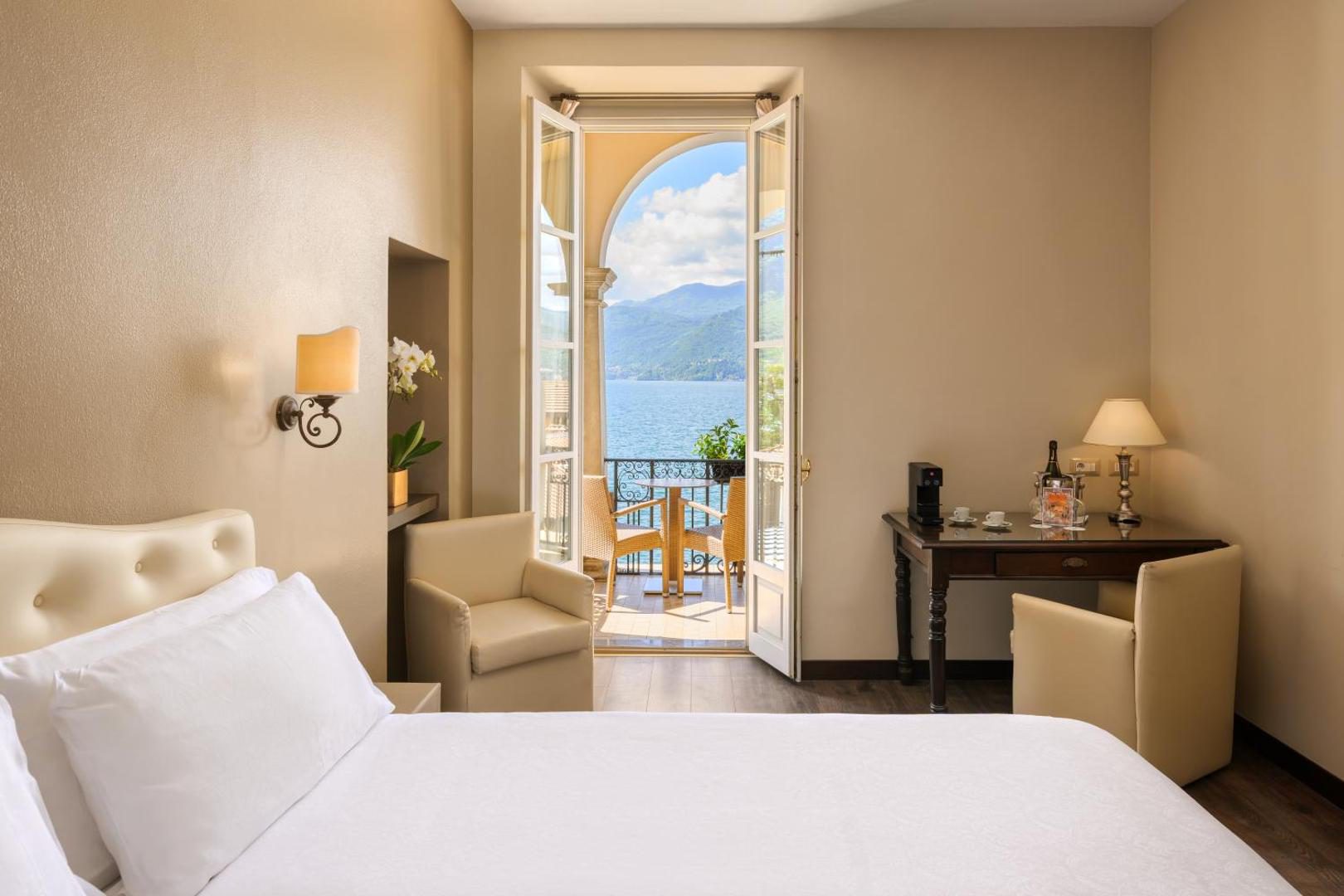 Hotel Royal Victoria Varenna – camera eleganta cu pat alb, fotoliu crem si usa arcuita cu balcon cu vedere la Lacul Como si munti, Varenna Hotel Royal Victoria Varenna – camera eleganta cu pat alb, fotoliu crem si usa arcuita cu balcon cu vedere la Lacul Como si munti, Varenna