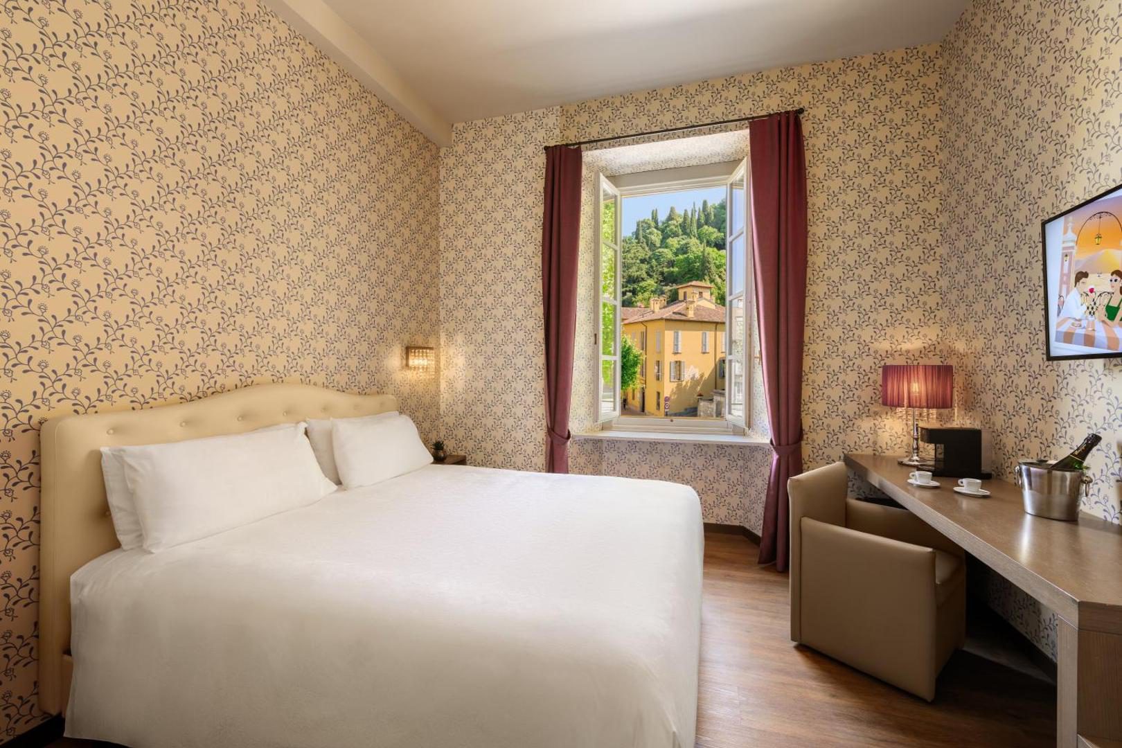 Hotel Royal Victoria Varenna – camera clasica cu tapet floral, pat dublu alb, birou si fereastra cu vedere la orasul Varenna, Lacul Como Hotel Royal Victoria Varenna – camera clasica cu tapet floral, pat dublu alb, birou si fereastra cu vedere la orasul Varenna, Lacul Como