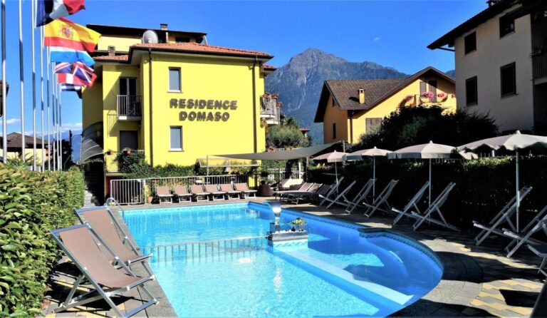Residence Domaso – piscina exterioara sezoniera cu sezlonguri si umbrele, cladire galbena cu steaguri si munti in fundal, Domaso