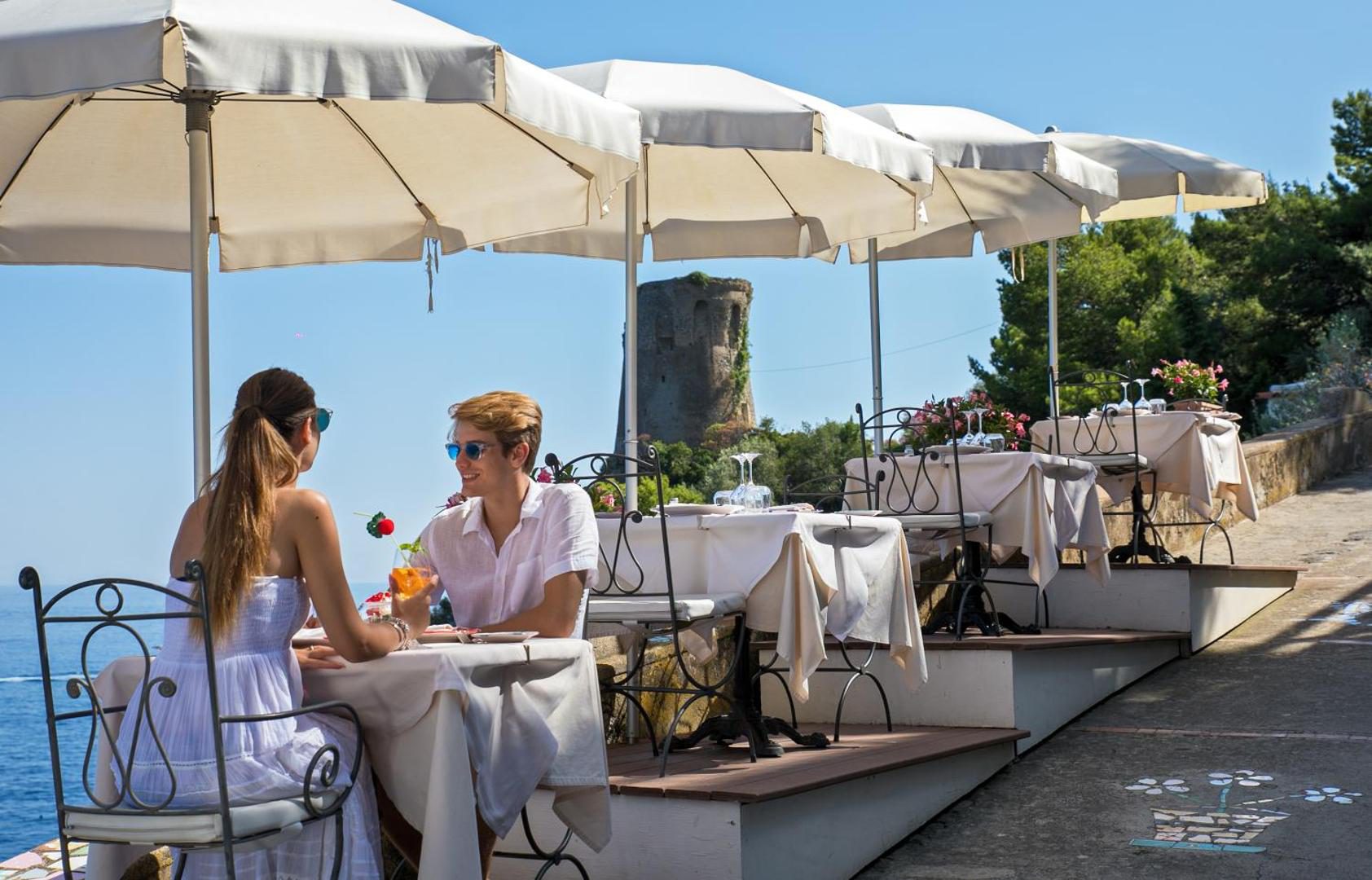 Hotel Onda Verde Praiano – terasa restaurant cu umbrele albe si vedere la turnul medieval de coasta Hotel Onda Verde Praiano – terasa restaurant cu umbrele albe si vedere la turnul medieval de coasta