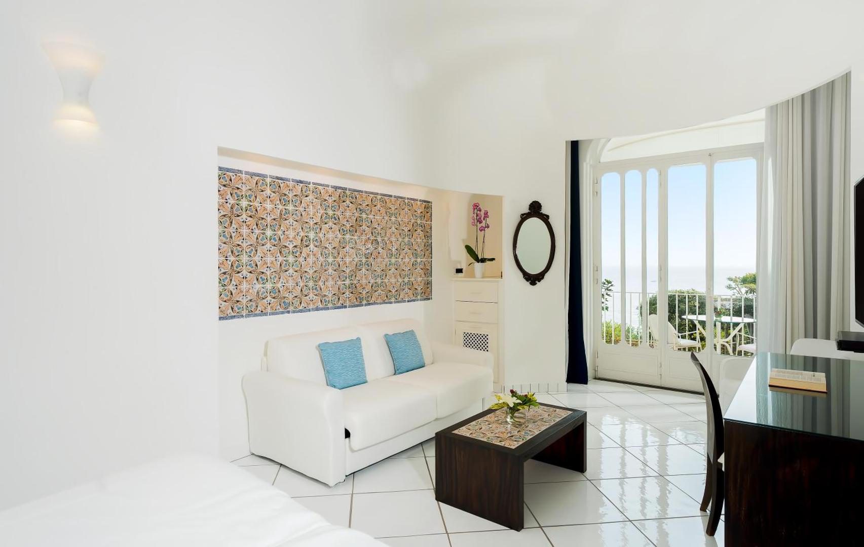 Hotel Santa Caterina Amalfi – living cu faianta pictata manual, canapea alba si vedere la mare prin usa balconului Hotel Santa Caterina Amalfi – living cu faianta pictata manual, canapea alba si vedere la mare prin usa balconului