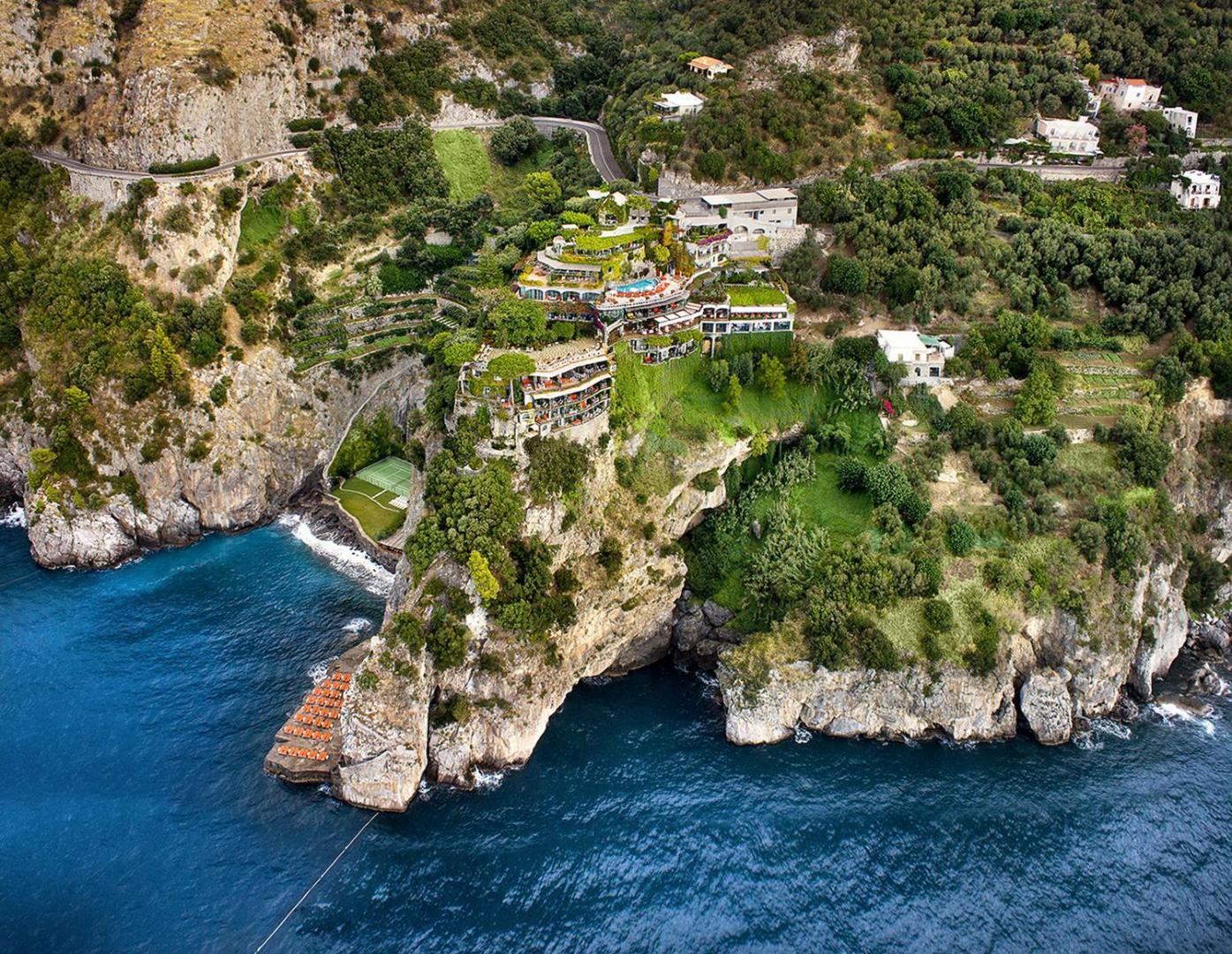 Il San Pietro di Positano – vedere aeriana a hotelului construit in terase pe stanca, cu piscina exterioara, teren de tenis si plaja privata la baza stancii, Positano