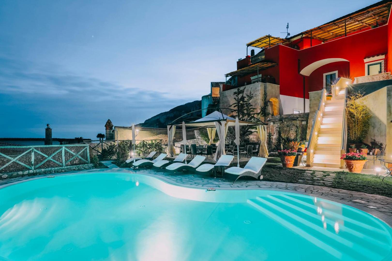 Palazzo Pascal Scala – piscina infinita in amurg, cu sezlonguri si baldachine, vedere la palat si munte Palazzo Pascal Scala – piscina infinita in amurg, cu sezlonguri si baldachine, vedere la palat si munte