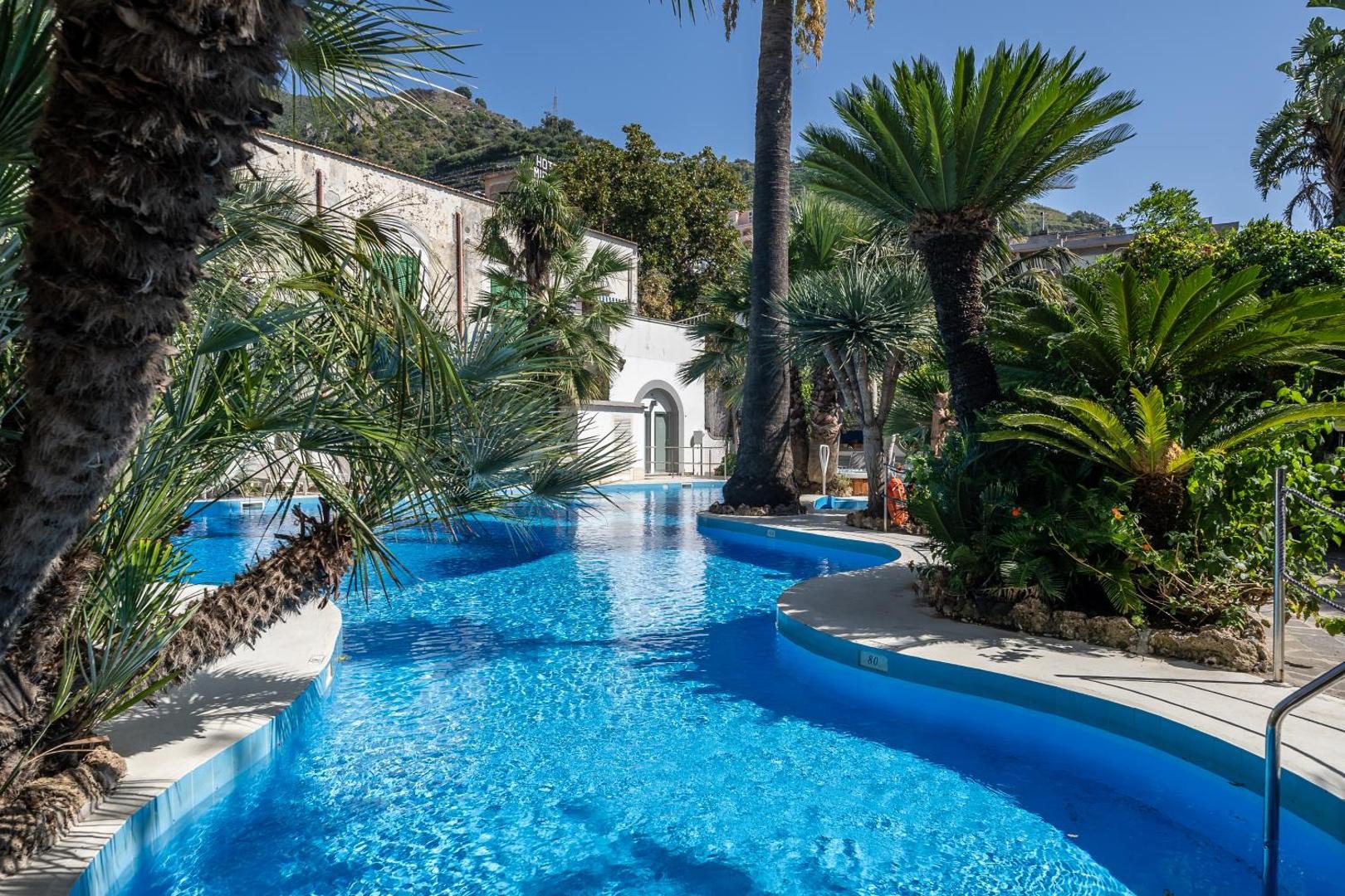 Reginna Palace Hotel Maiori – piscina cu forma sinuoasa inconjurata de palmieri si plante tropicale Reginna Palace Hotel Maiori – piscina cu forma sinuoasa inconjurata de palmieri si plante tropicale